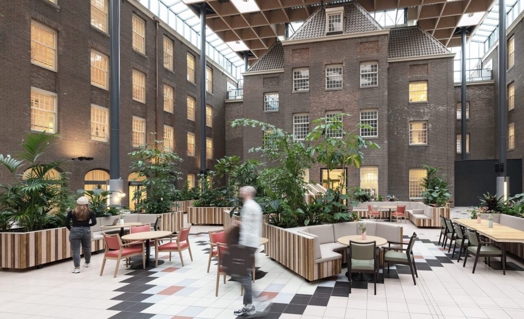 A spacious office atrium in Dr. Sarphatihuis, Amsterdam, featuring historic brick architecture, modern wooden seating, lush greenery, and a glass ceiling allowing natural light.