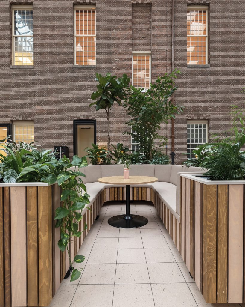 Biophilic office lounge with a built-in wooden bench, round meeting table, and lush greenery, set against an exposed brick wall with large windows