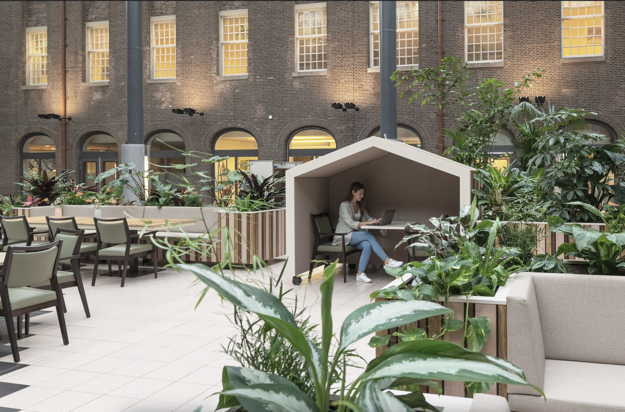 A modern indoor workspace designed by MOSS, featuring lush biophilic elements, flexible seating, and a cozy, house-shaped acoustic booth where a woman is working on her laptop. The space integrates greenery, natural light, and soft materials to create a sustainable and inspiring work environment.