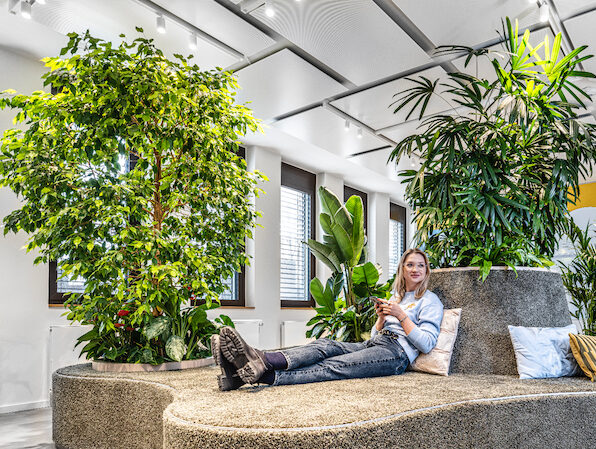 Medewerker ontspant op een organisch gevormd lounge-eiland, omringd door grote indoor bomen en tropische planten in het JetBrains-kantoor in München.