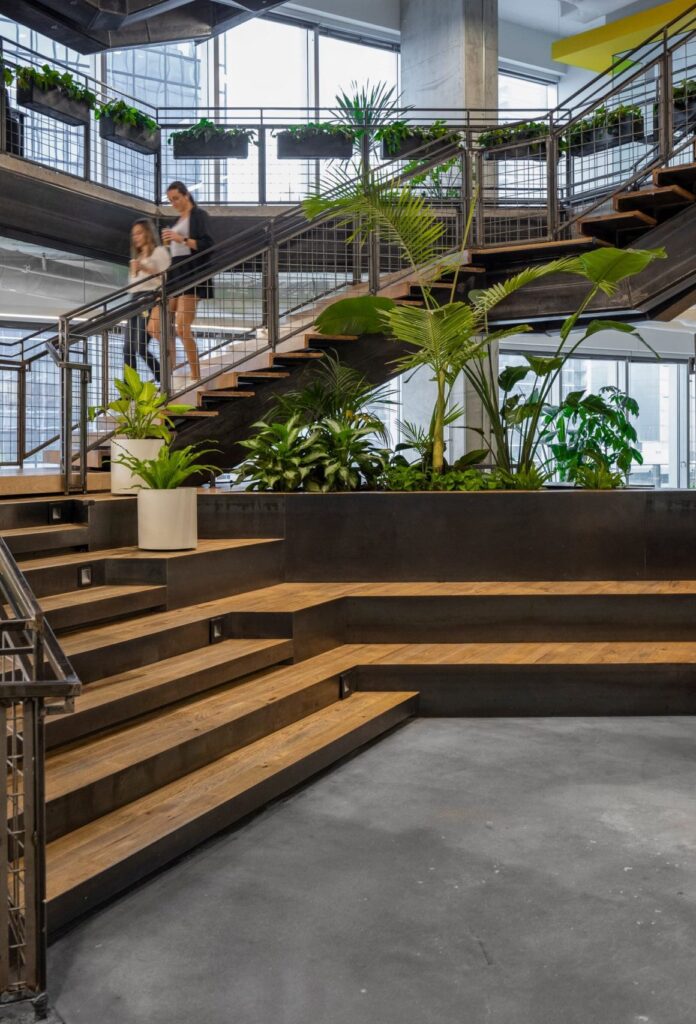 Twee medewerkers lopen een industriële trap af in het Miro-kantoor in Austin, omringd door grote tropische planten en een houten tribunetrap met geïntegreerde beplanting.