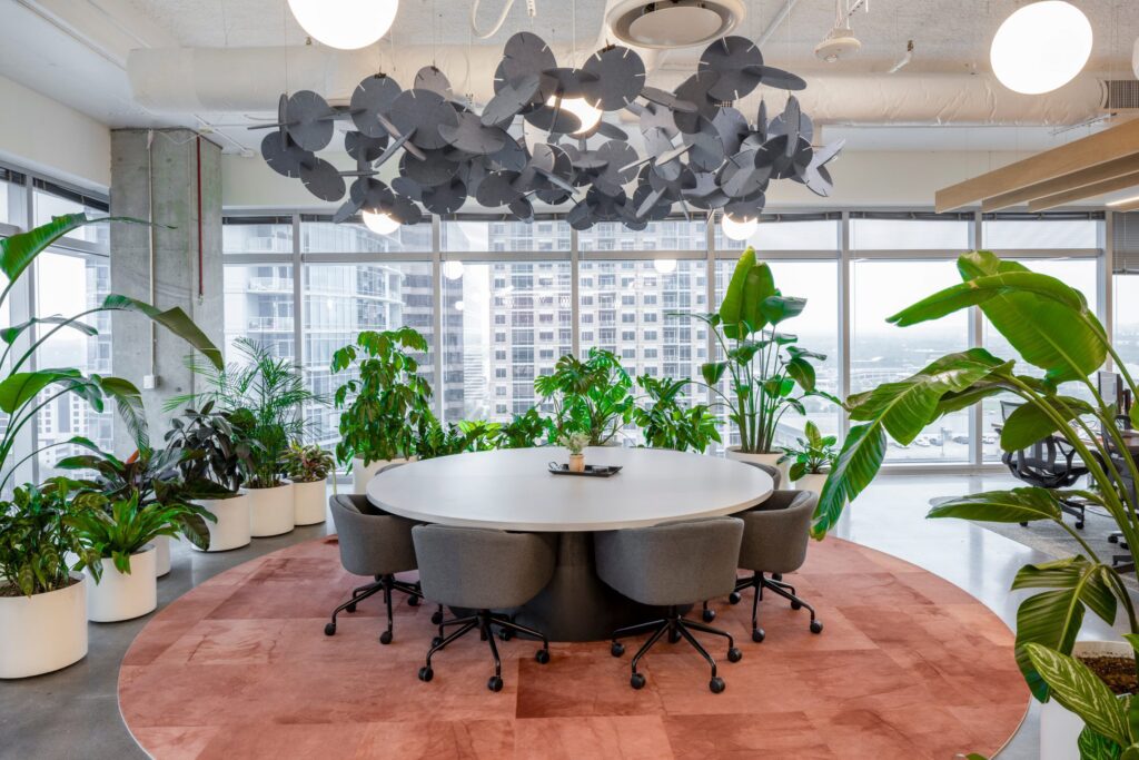 Overlegruimte in het Miro-kantoor in Austin met een grote ronde tafel, omringd door witte plantenbakken vol tropische planten en grote ramen met uitzicht op de stad.