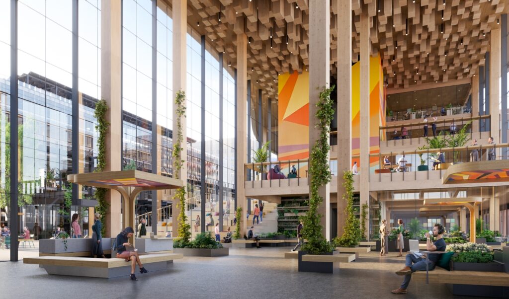 Impressie van het atrium van District West Amsterdam met hoge houten kolommen, verticale beplanting, veel daglicht en informele werk- en ontmoetingsplekken.