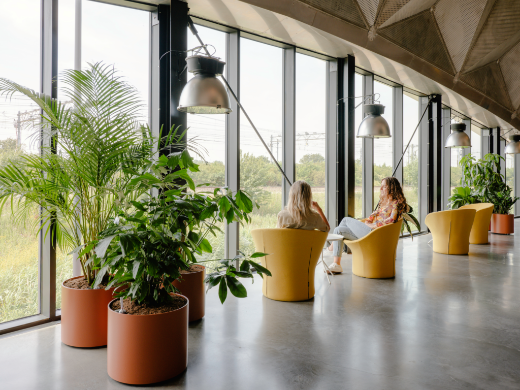 Indoor tropical plants by MOSS frame a lounge area beside floor-to-ceiling windows inside Circa Amsterdam, creating a calming biophilic environment overlooking the surrounding landscape.