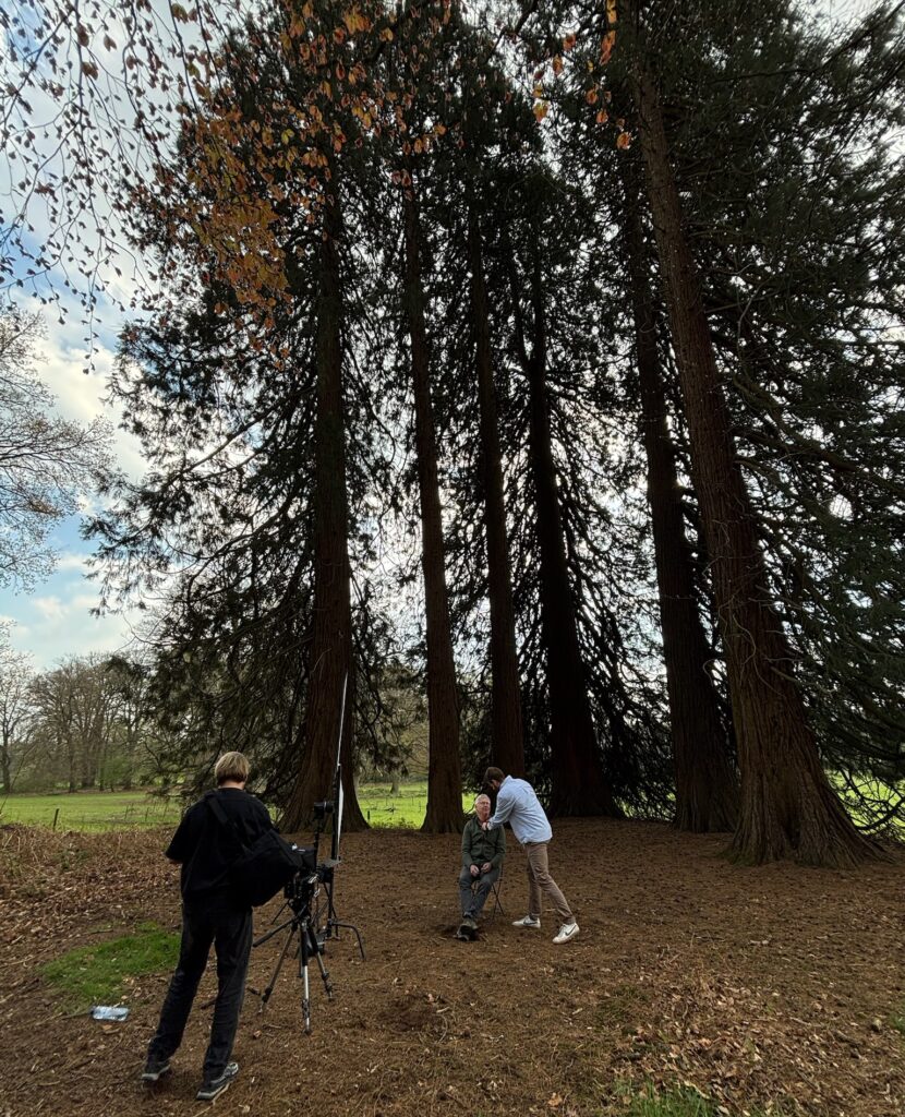 Film crew interviewing ecologist in dense forest with tall conifer trees, showcasing natural woodland structure and ecological design