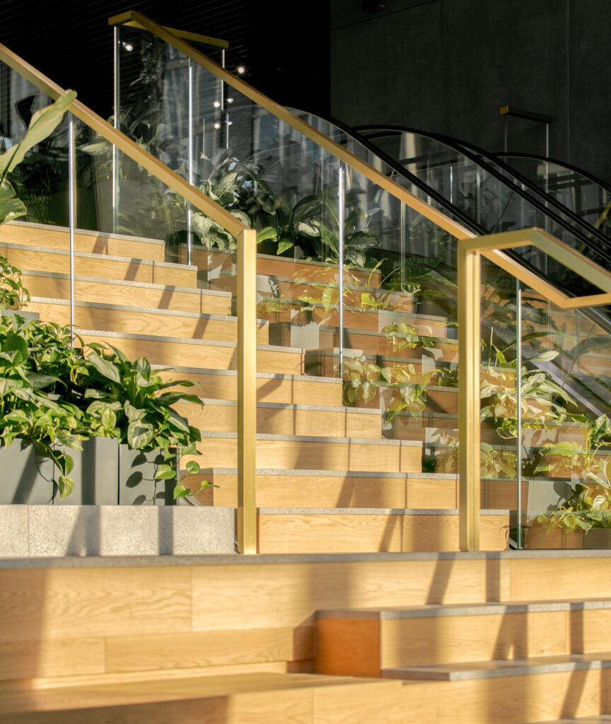 Sunlit wooden stairs bordered by leafy planters, glass balustrades and gold-toned handrails at The Bridge in Warsaw.